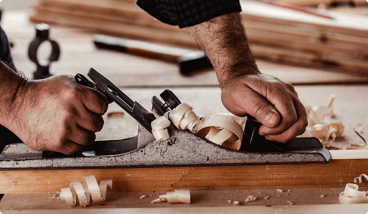 Carpenter hand-planing a timber board, wood shavings curling off the blade
